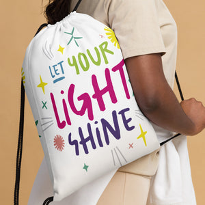 A woman holding a white drawstring bag. The drawstring bag has the words "Let Your Light Shine" with sketchy illustrations around the words. 