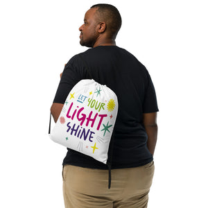 A man with a white drawstring bag on his back. The drawstring bag has the words "Let Your Light Shine" with sketchy illustrations around the words. 