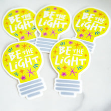 Load image into Gallery viewer, Die cut vinyl stickers with the words &quot;be the light&quot; inside an illustrated lightbulb.