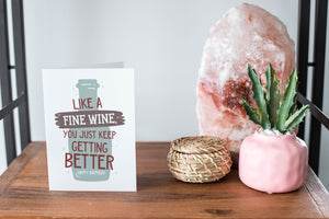 A card on a wood tabletop and on the right side of the card is a woven basket, a pink plant pot with a cactus in it and a pink crystal rock. The card features the words "Like a fine wine, you keep getting better, Happy Birthday!” with an illustration of a wine bottle behind the words.