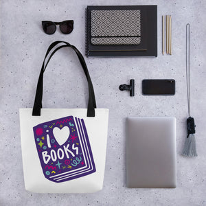 A white tote bag with black handles on a grey tabletop with a laptop and other office supplies. The tote bag has a design with the words 'I love books' with the word love shown as a heart. The words are inside an illustrated book. 