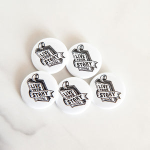 Buttons on a white tabletop. The buttons feature word art illustrations with the words "Live Your Story" inside an illustrated book.  