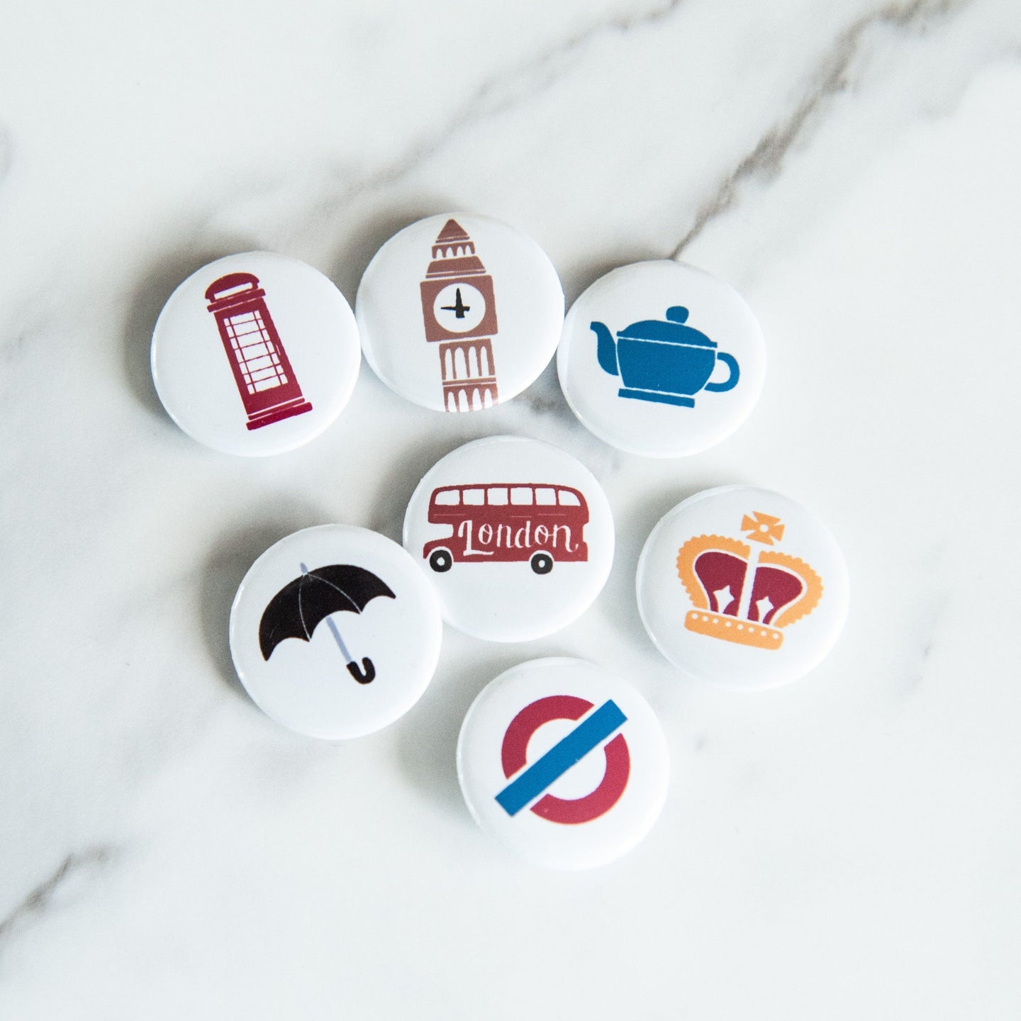 A set of seven buttons on white tabletop. The buttons features illustrations of a red telephone booth, Big Ben, a teapot, a royal crown, a double decker bus, a black umbrella and the London underground/tube logo. 