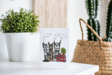 Load image into Gallery viewer, A greeting card is featured on a white tabletop with a white planter in the background with a green plant. There’s a woven basket in the background with a cactus inside. The card features a design with illustrated London houses, a black taxi cab and a red double decker bus.