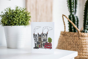 A greeting card is featured on a white tabletop with a white planter in the background with a green plant. There’s a woven basket in the background with a cactus inside. The card features a design with illustrated London houses, a black taxi cab and a red double decker bus.