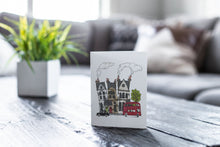 Load image into Gallery viewer, A greeting card featured on a black, wood coffee table. There’s a white planter in the background with a green plant. There’s also a gray sofa in the background with a white pillow. The card features a design with illustrated London houses, a black taxi cab and a red double decker bus.