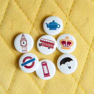 A set of seven buttons on a yellow background. The buttons features illustrations of a red telephone booth, Big Ben, a teapot, a royal crown, a double decker bus, a black umbrella and the London underground/tube logo. 