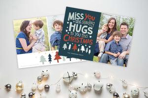A photo of a Christmas card showing the front and back of the card laying on a white surface. To the bottom of the cards are silver and white small ornaments. The front of the card features a photo on the right side and on the left side are the words “ Miss you and sending hugs to you this Christmas” with illustrated trees below the words. The back of the card features two photos with illustrated trees at the bottom.