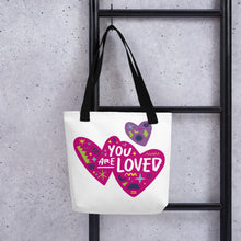 Load image into Gallery viewer, A white tote bag with black handles handing on a black ladder. The bag features a design with the words &#39;you are loved&#39; inside two illustrated pink hearts with a smaller, purple heart above. 