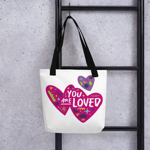 A white tote bag with black handles handing on a black ladder. The bag features a design with the words 'you are loved' inside two illustrated pink hearts with a smaller, purple heart above. 