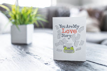 Load image into Gallery viewer, A greeting card featured on a black, wood coffee table. There’s a white planter in the background with a green plant. There’s also a gray sofa in the background with a white pillow. The card features the words “You are my love story” with an illustrated typewriter and scattered papers.