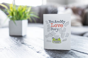 A greeting card featured on a black, wood coffee table. There’s a white planter in the background with a green plant. There’s also a gray sofa in the background with a white pillow. The card features the words “You are my love story” with an illustrated typewriter and scattered papers.