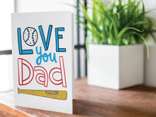 Load image into Gallery viewer, A greeting card is featured on a wood coffee table with a green plant in a white planter in the background. The card features the words  “Love You Dad” with an illustrated baseball as the “O” of love and a baseball bat featured at the bottom of the words. 