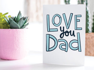 A greeting card featured standing up on a white tabletop with a pink plant pot in the background and some succulents in the pot. There’s a woven basket in the background with a cactus inside. The card features the words “Love You Dad” in simple typography. 