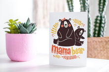 Load image into Gallery viewer, A greeting card featured standing up on a white tabletop with a pink plant pot in the background and some succulents in the pot. There’s a woven basket in the background with a cactus inside. The card features the words “Mama Bear” with an illustrated mama bear and baby bear. 