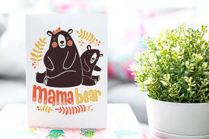A greeting card is on a table top with a gift in pink wrapping paper. Next to the gift is a white plant pot with a green plant. The card features the words “Mama Bear” with an illustrated mama bear and baby bear.