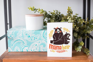 A greeting card is on a table top with a present in blue wrapping paper in the background. On top of the present is a candle and some greenery from a plant too. The card features the words “Mama Bear” with an illustrated mama bear and baby bear. 