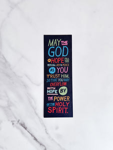 A bookmark laying on a marble tabletop with a black background on the bookmark and colorful letters with the words "May the God of hope fill you with all joy & peace as you trust Him, so that you may overflow with hope by the power of the Holy Spirit."