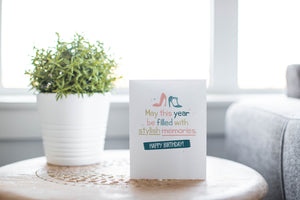 A greeting card is featured on a wood coffee table with a green plant in a white planter in the background. The card features the words “May This Year be filled with stylish memories, Happy Birthday: with illustrated heels above the words. 