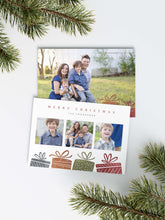 Load image into Gallery viewer, A photo of a Christmas card showing the front and back of the card laying on a white surface. Around the two sides of the card are pine needles. The front of the card features three photos with illustrated gifts on the bottom. On top of the photos the words “Merry Christmas” is featured with a family name below which you can edit. The back of the card features one photo with some more illustrated gifts.
