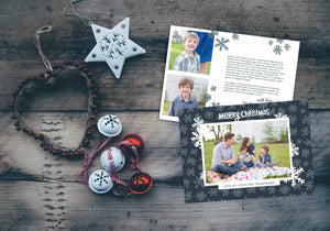 A photo of a Christmas card showing the front and back of the card laying on a wood surface. To the left of the cards are some ornaments. The front of the card features a photo with a frame around it with illustrated snowflakes. Above the photo reads “Merry Christmas” and below the photo you can add your family name. The back of the card features two photos, illustrated snowflakes and a place to add a family update.
