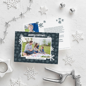 A photo of a double-sided Christmas card showing the front and back of the card laying on a white surface. Around the two sides of the card are surrounded with Christmas items. The front of the card features a photo with a frame around it with illustrated snowflakes. Above the photo reads “Merry Christmas” and below the photo you can add your family name. The back of the card features two photos, illustrated snowflakes and a place to add a family update. 