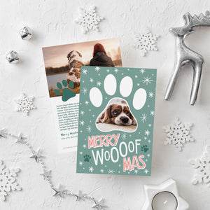 A photo of a double-sided Christmas card showing the front and back of the card laying on a white surface. Around the two sides of the card are surrounded with Christmas items. The front of the card features a photo inside a paw shaped frame. Around the photo are white stars and below are the words “Merry Woofmas.” The back of the card features one photo with a dog paw illustration and space to add an update.