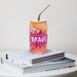 A photo of a glass can tumbler on a coffee table on top of books. The glass tumbler has a bamboo lid and silver metal straw. The design on the glass tumbler says the word "brave" inside a hot pink star with illustrated sketchy elements around the word. 