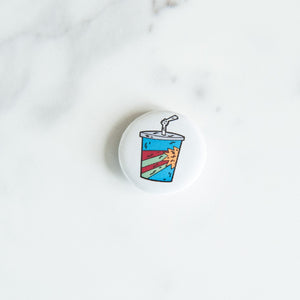 A button pin on a white tabletop. The pin features an illustration of a milkshake cup.