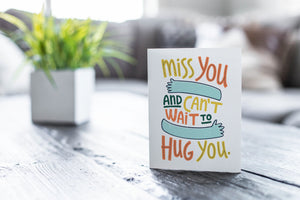 A greeting card featured on a black, wood coffee table. There’s a white planter in the background with a green plant. There’s also a gray sofa in the background with a white pillow. The card features the words “Miss you and can’t wait to hug you.”
