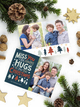 Load image into Gallery viewer, A photo of a two-sided Christmas card showing the front of the card on top of a brown wrapped gift on a white tabletop. Around the gift are pine needles, pinecones and wood star ornaments. The front of the card features a photo on the right side and on the left side are the words “ Miss you and sending hugs to you this Christmas” with illustrated trees below the words. The back of the card features two photos with illustrated trees at the bottom.