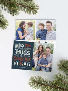 A photo of a Christmas card showing the front and back of the card laying on a white surface. Around the two sides of the card are pine needles. The front of the card features a photo on the right side and on the left side are the words “ Miss you and sending hugs to you this Christmas” with illustrated trees below the words. The back of the card features two photos with illustrated trees at the bottom.