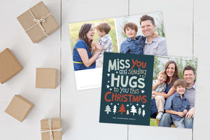A photo of a Christmas card showing the front and back of the card laying on a white surface. Brown wrapped small gifts are to the left of the cards. The front of the card features a photo on the right side and on the left side are the words “ Miss you and sending hugs to you this Christmas” with illustrated trees below the words. The back of the card features two photos with illustrated trees at the bottom.