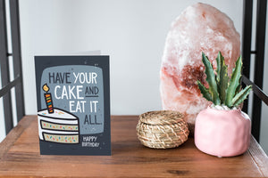 A card on a wood tabletop and on the right side of the card is a woven basket, a pink plant pot with a cactus in it and a pink crystal rock. The card features the words “Have Your Cake and Eat it All Happy Birthday” with an illustrated piece of cake with a candle in it.