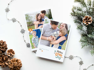 A photo of a double-sided Christmas card showing the front and back of the card laying on a white surface. Around the two sides of the card are pine cones, pine needles and a string of silver snowflake garland. The front of the card features a photo with the words “Peace Love joy” to the bottom right and space to the bottom left to put your family names. The words feature some modern illustrated flowers around it. The back of the card features three photos with modern illustrated flowers.