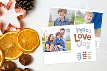 Load image into Gallery viewer, A photo of a Christmas card showing the front and back of the card laying on a white surface. Left of the card is a cookie cutter, pinecone, nuts and dried oranges. The front of the card features a photo on the left side with the words “Peace Love Joy” with modern illustrated leaves below. The back of the card features two photos with the modern illustrated leaves. 