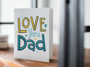 A card on a wood tabletop with an object in the background that is out of focus. The card features the words “Love you Dad” with small stars around the letters. 