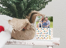 Load image into Gallery viewer, A photo of a one-sided Christmas card showing the front of the card standing up next to the bottom of a small Christmas tree with the base wrapped in burlap. The photo card features one photo with illustrated modern Christmas trees below. The words “Peace Love Joy” are to the left of the tree pattern. 