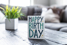 Load image into Gallery viewer, A greeting card is featured on a wood coffee table with a green plant in a white planter in the background. The card features the words “Happy birthday” with blue letters featured on a white background. 