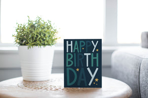 A greeting card is featured on a wood coffee table with a green plant in a white planter in the background. The card features the words “happy birthday” with stars surrounding the letters.