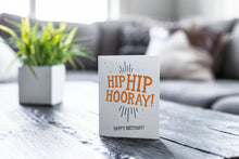 Load image into Gallery viewer, A greeting card featured on a black, wood coffee table. There’s a white planter in the background with a green plant. There’s also a gray sofa in the background with a white pillow. The card features the words “Hip Hip Hooray! Happy Birthday!” The words &quot;digital download&quot; are featured in a circle on top of the image.