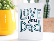 Load image into Gallery viewer, A greeting card is on a table top with a yellow plant pot and a green plant inside. The card features the words “Love You Dad” in simple typography. 