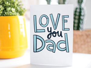 A greeting card is on a table top with a yellow plant pot and a green plant inside. The card features the words “Love You Dad” in simple typography. 