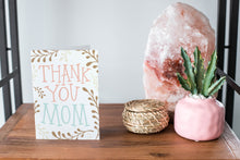 Load image into Gallery viewer, A card on a wood tabletop and on the right side of the card is a woven basket, a pink plant pot with a cactus in it and a pink crystal rock. The card features the words “Thank You Mom” with illustrated plant leaves.