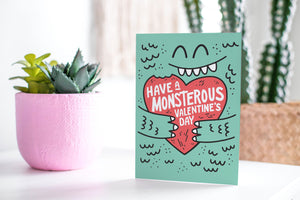 A greeting card featured standing up on a white tabletop with a pink plant pot in the background and some succulents in the pot. There’s a woven basket in the background with a cactus inside. The card features the words “Have a monstrous Valentine’s Day” with an illustrated monster holding a heart.