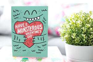 A greeting card is on a table top with a gift in pink wrapping paper. Next to the gift is a white plant pot with a green plant. The card features the words “Have a monstrous Valentine’s Day” with an illustrated monster holding a heart.