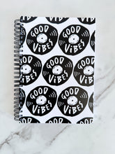 Load image into Gallery viewer, A spiral notebook is on a tabletop. The white spiral notebook has black spiral binding. The pattern design features the words &quot;good vibes&quot; inside an illustrated vinyl record.  