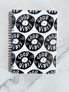 A spiral notebook is on a tabletop. The white spiral notebook has black spiral binding. The pattern design features the words "good vibes" inside an illustrated vinyl record.  