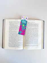 Load image into Gallery viewer, A bookmark placed inside a book. The bookmark features the words &quot;wild and free&quot; inside illustrated snow-capped mountains with illustrated trees. 