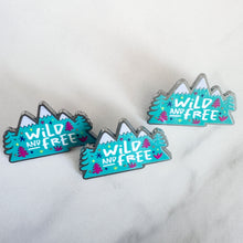 Load image into Gallery viewer, A top view of three enamel pins featuring the words &quot;wild and free&quot; inside illustrated mountains with illustrated trees. 